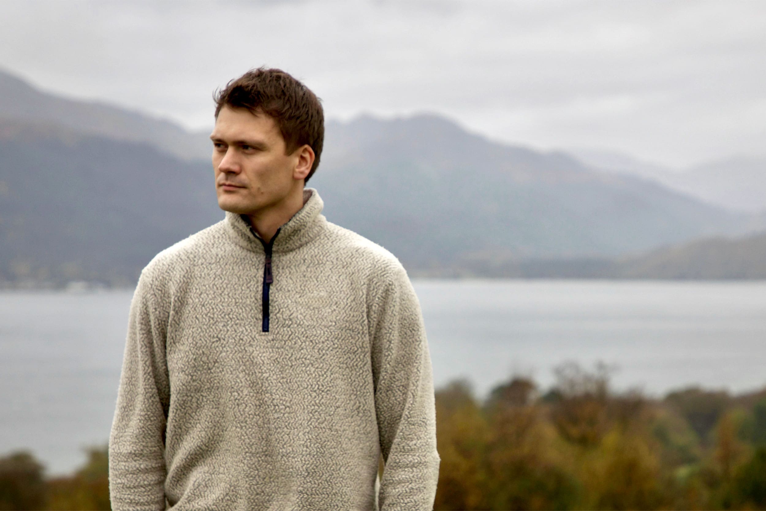 A man in a cream-coloured woollen top stands in front of a large lake or loch with hills in the backgroud.