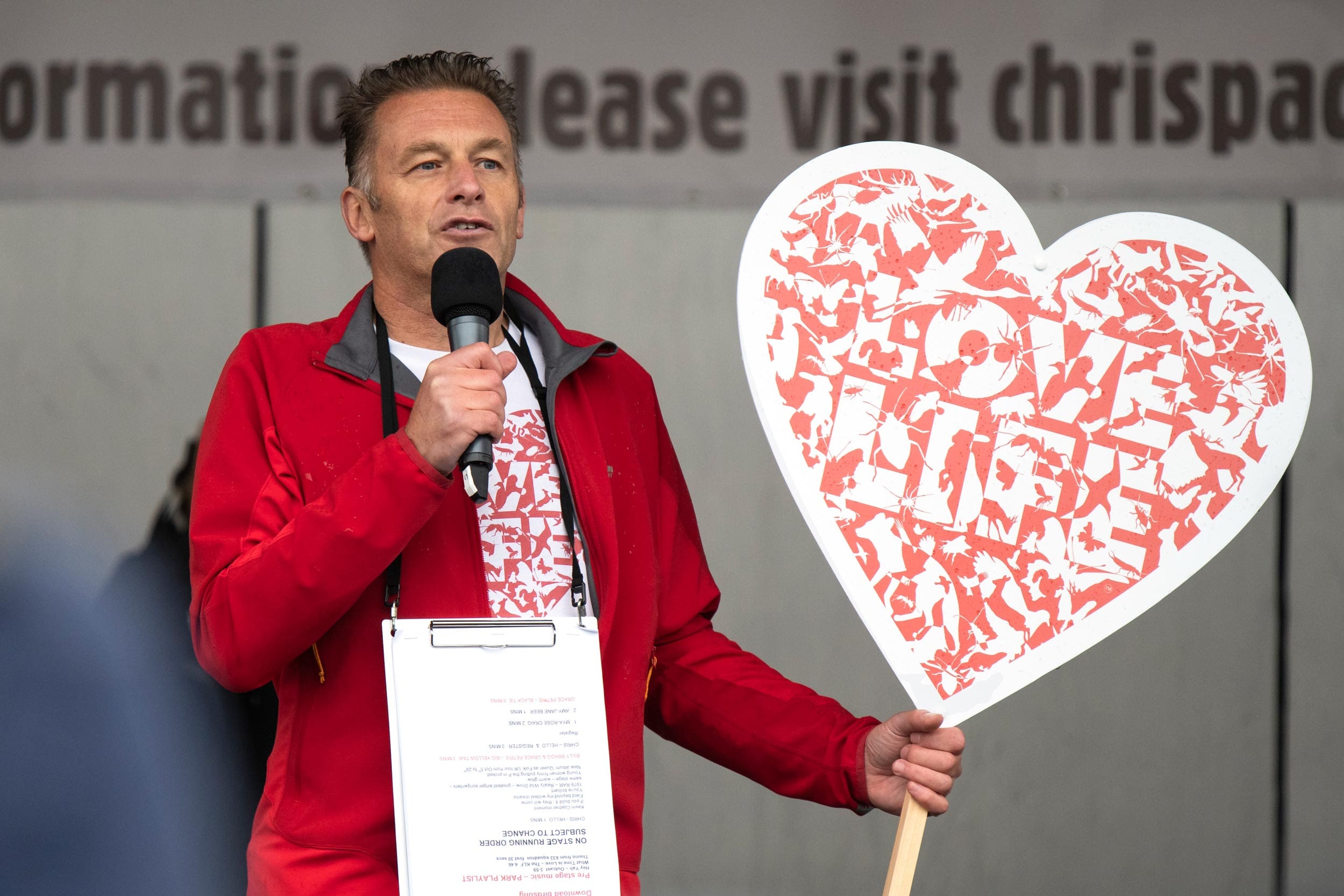 Chris Packham is speaking into a microphone and holding a heart-shaped sign that reads 'Love Life' with graphics of animals on it