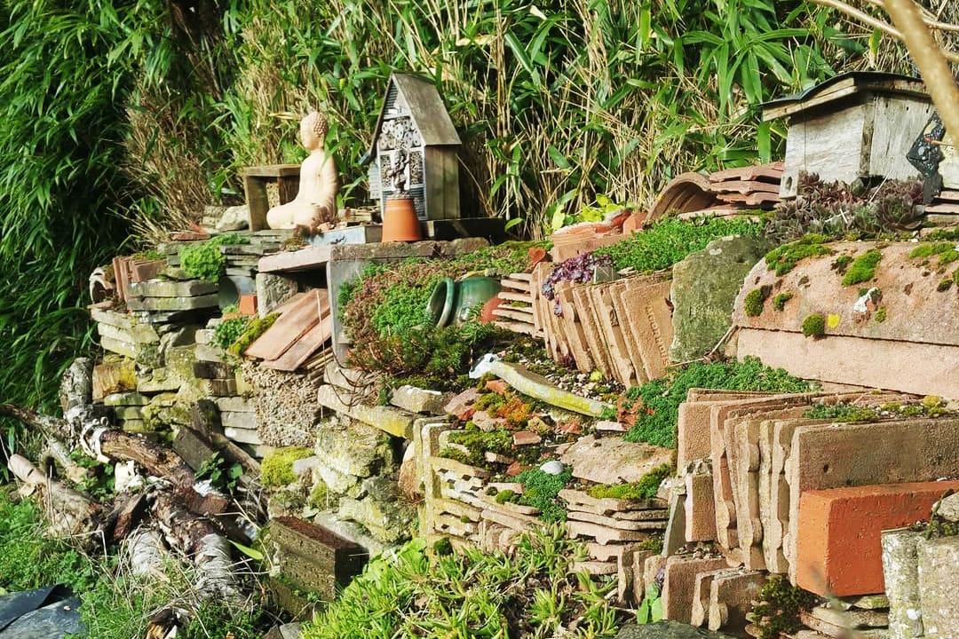 A wall constructed of different types of roof tiles, stacked in sections, with moss and plants growing out and over them