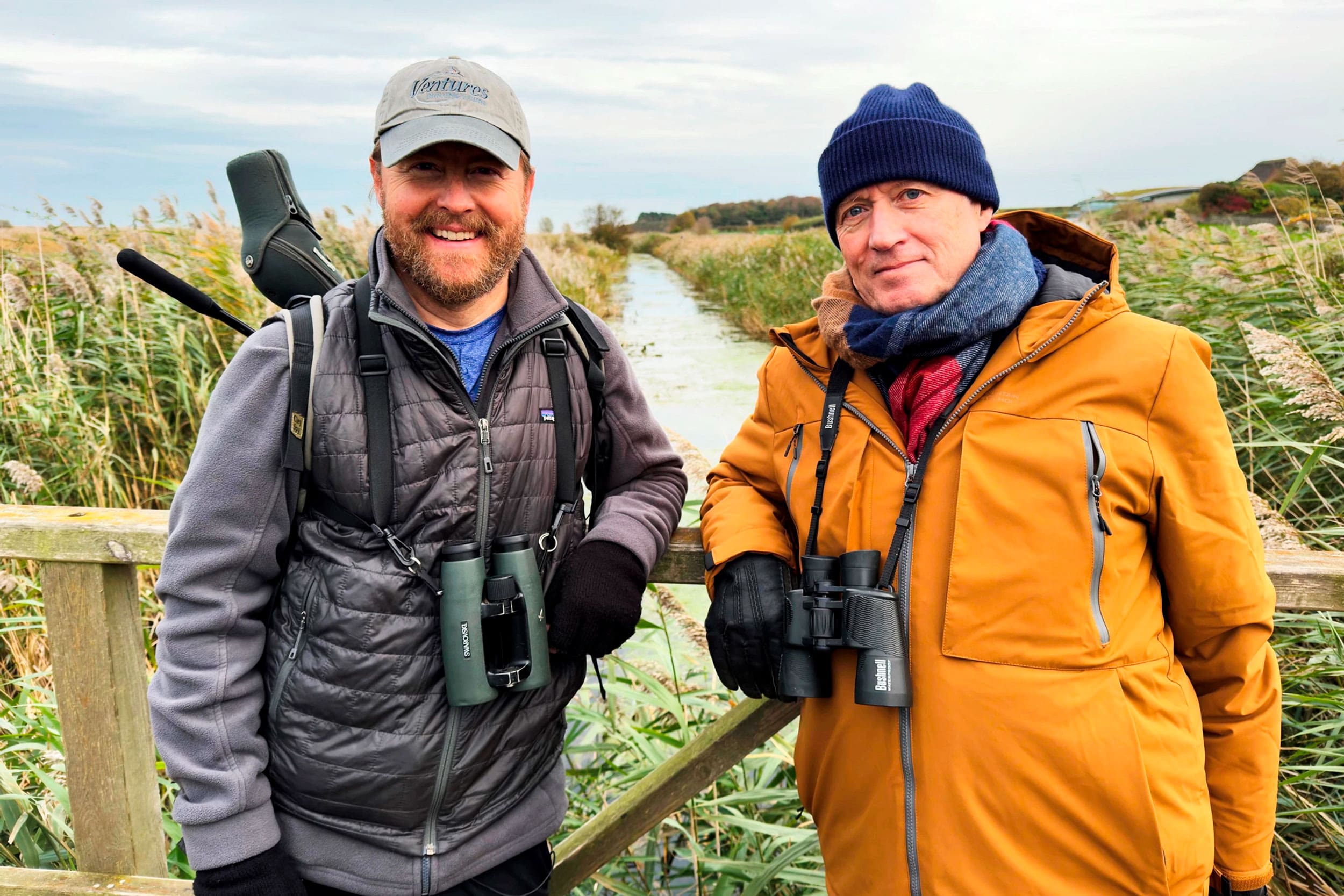 Sam and Ade lean against the side of a small wooden bridge, over a river running through a wetland landscape. They both hold their binoculars